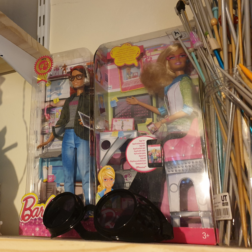 Two Barbie dolls still in their boxes, next to a container of knitting needles