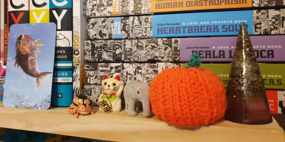 A shelf with graphic novels. In front of the books is a card with a magikarp on it, a small toy catbus, a small lucky cat, a small elephant, a knitted pumpkin, and a cone made from resin with glitter and rocks in it.