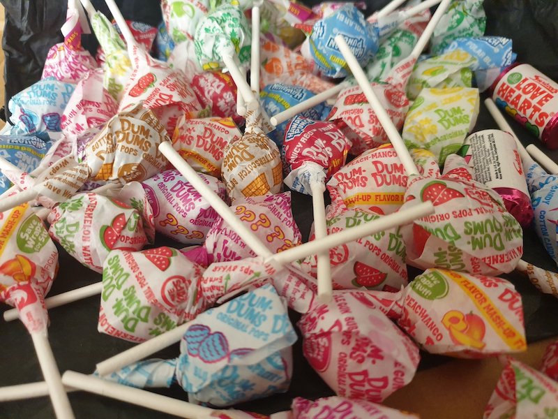 A collection of Dum-Dum Lollipops, scattered in a box, with a couple of rolls of Love Hearts hiding among them.